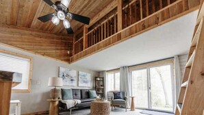 A spacious living room with a high ceiling, wooden beams, a ceiling fan, a sofa, a coffee table, a bookshelf, and a large sliding glass door. - Sunset Beach View on Otter Tail Lake (Otter Tail)