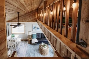 A loft-style room with a high ceiling, wooden beams, a ceiling fan, a sofa, a dining table with chairs, and a window with blinds. - Sunset Beach View on Otter Tail Lake (Otter Tail)
