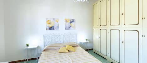A bedroom with a bed, two bedside tables, a chandelier, a wardrobe, and two wall-mounted paintings.