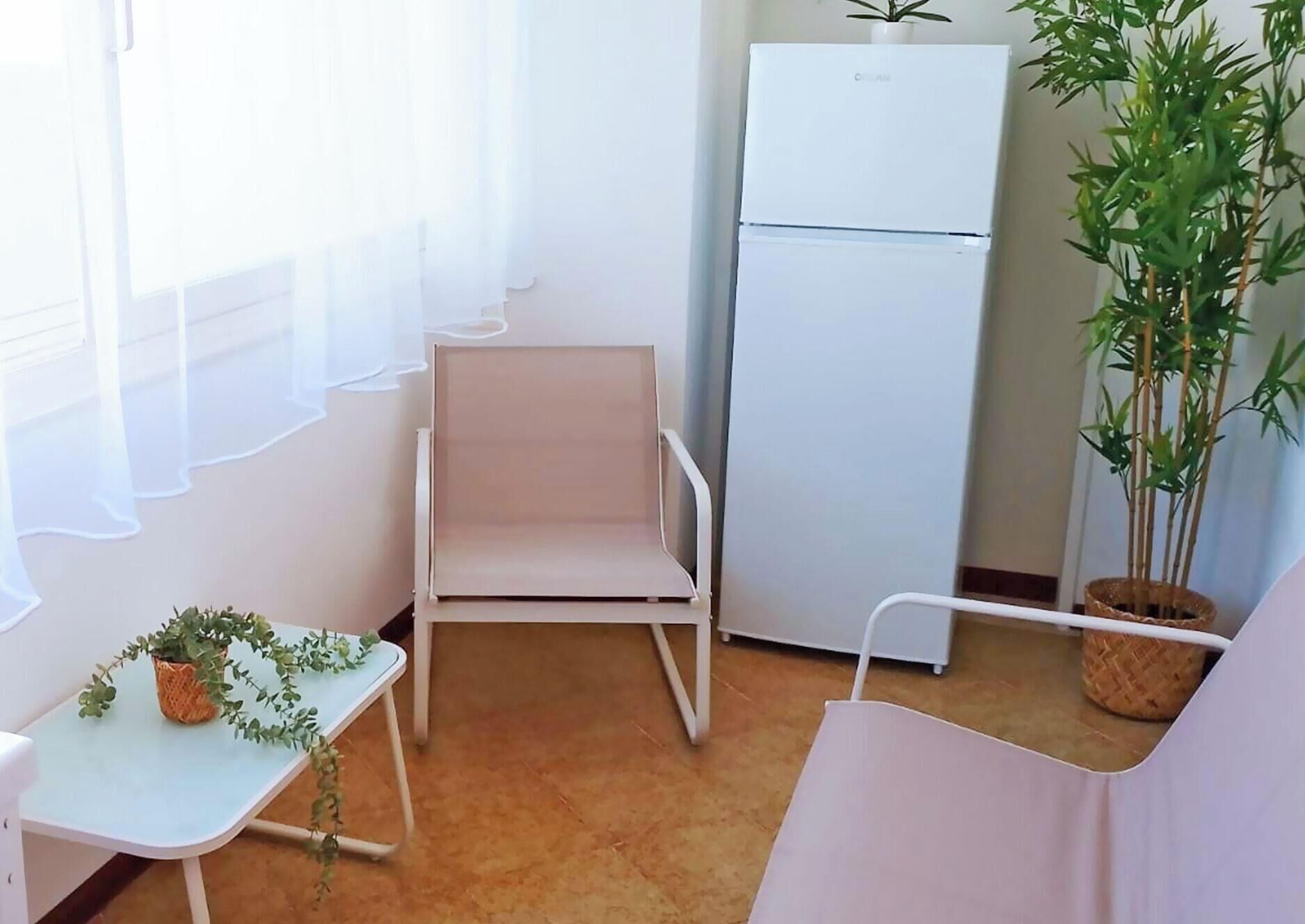 A compact room with a white chair, a small table with a plant, a white refrigerator, and a potted plant.