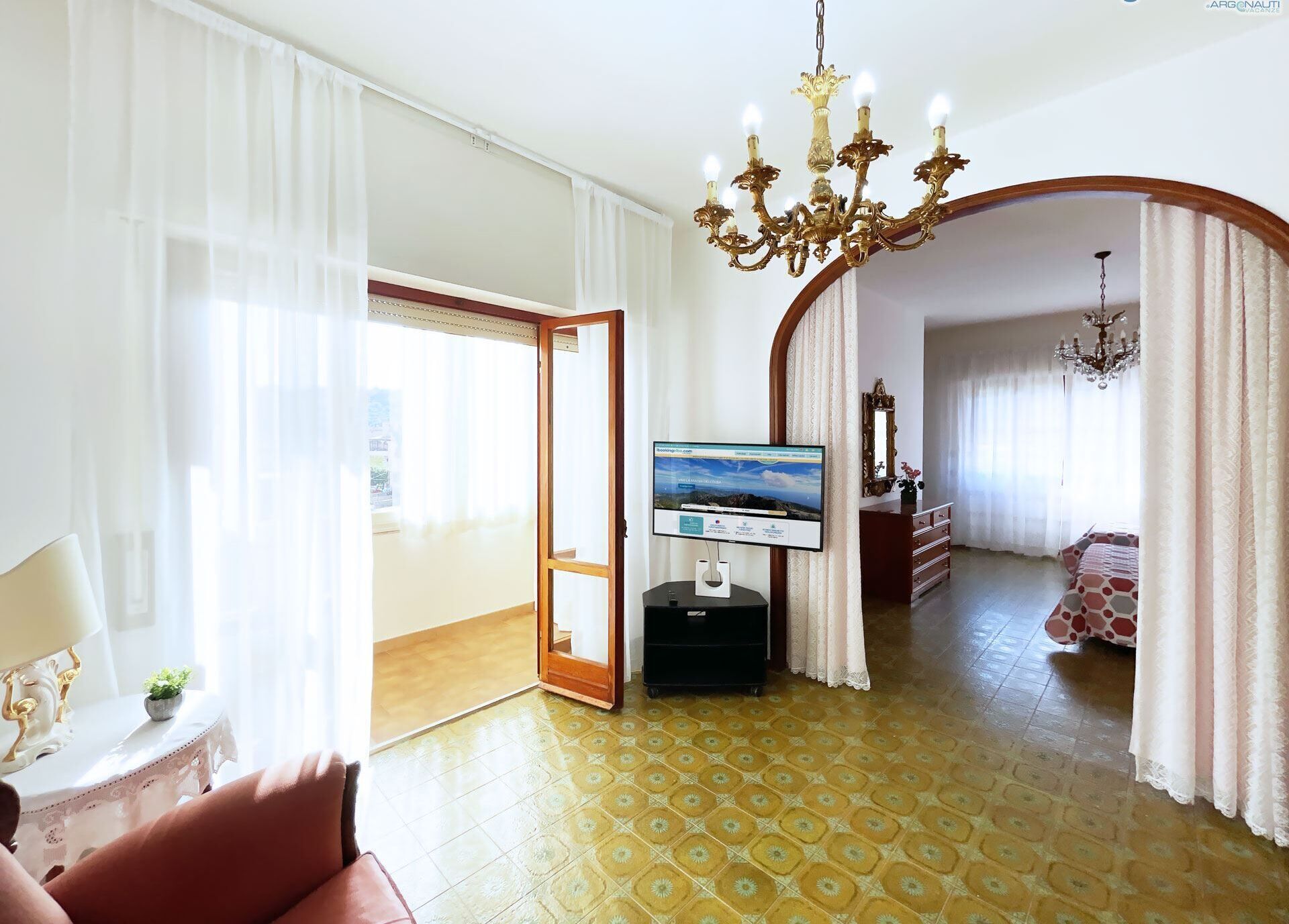 A hotel room with a television, a chandelier, a bed with patterned bedding, a wooden dresser, and a large window with sheer curtains.