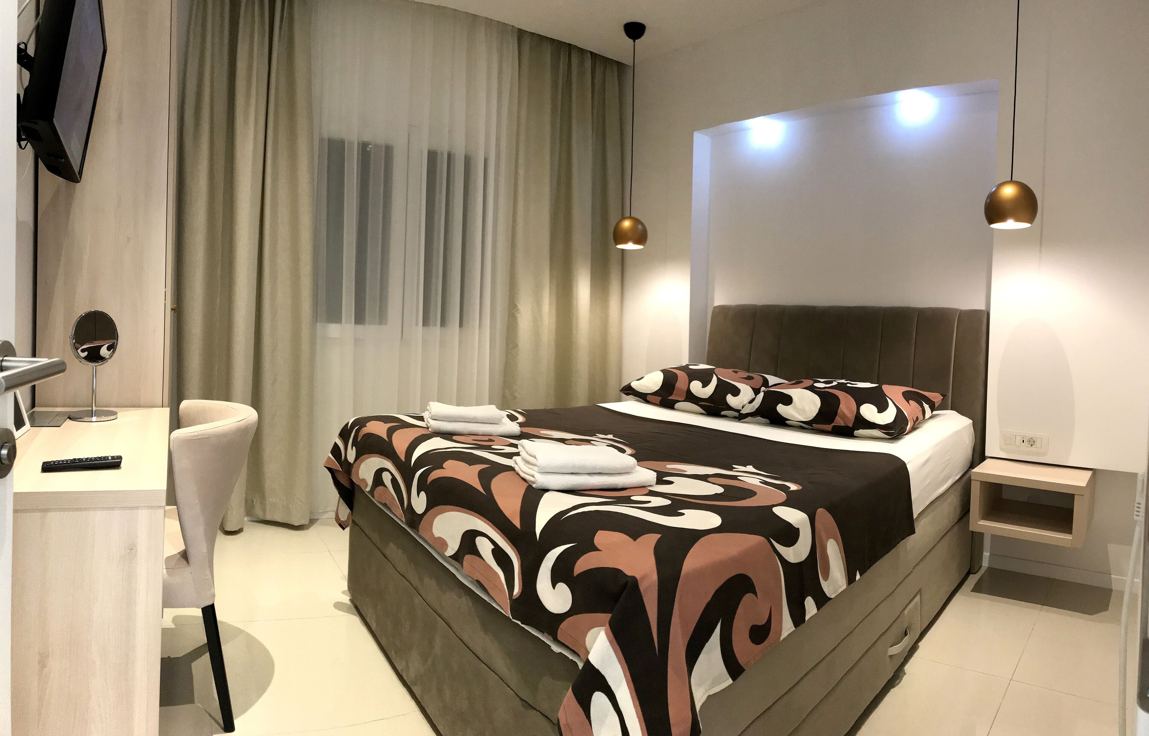A modern hotel room with a large bed, a desk with a chair, a TV, and a bedside table.