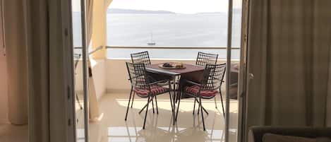 A balcony with a table and chairs, a view of the sea, and a sailboat in the distance.