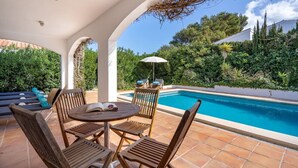 Un espace aménagé au bord de la piscine, avec une table et des chaises en bois rondes, un parasol blanc et une vue sur une piscine entourée d’une végétation luxuriante.