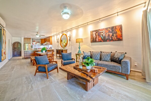 A spacious living area with a sofa, chairs, and a coffee table. There is a kitchen in the background with a dining table and chairs.