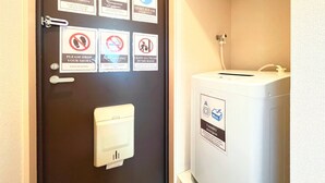 Porta di una camera d'albergo con diversi cartelli, tra cui il numero massimo di occupanti, una richiesta di silenzio, divieto di fumare e divieto di scarpe. Si vede anche una lavatrice con l'etichetta del detersivo.