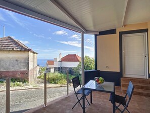 Een balkon met een glazen tafel en stoelen, een schaal fruit en uitzicht op huizen en de zee.