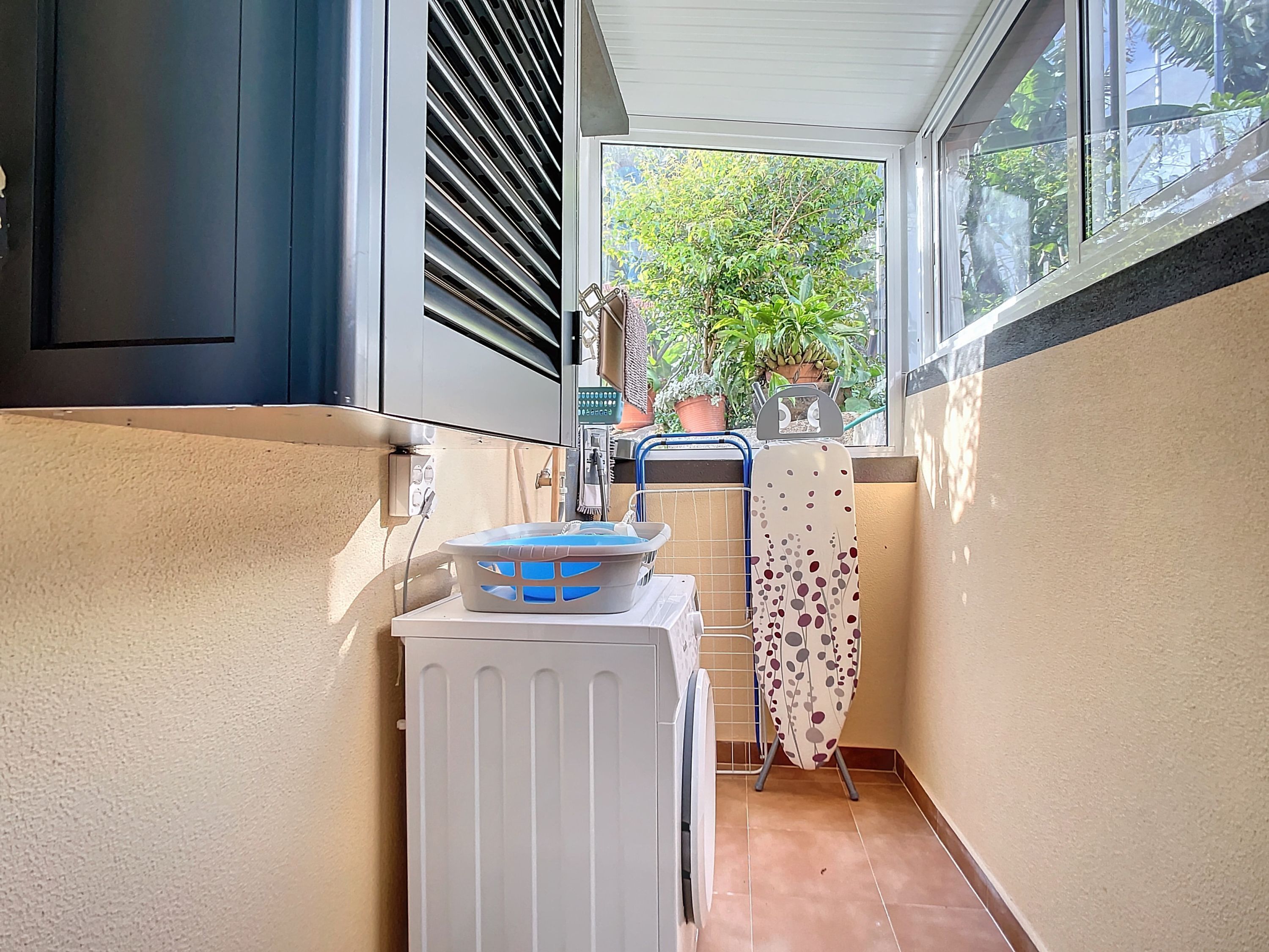 A narrow corridor with a washing machine, a drying rack, and a window with a view of greenery outside.