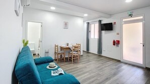 A modern hotel room with a blue sofa, a dining table with chairs, a TV mounted on the wall, and a door with a fire alarm. - Gomes House by Atlantic Holiday (Ponta do Sol)