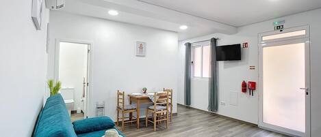 Ein modernes Hotelzimmer mit einem blauen Sofa, einem Esstisch mit Stühlen, einem an der Wand befestigten Fernseher und einer Tür mit Rauchmelder.