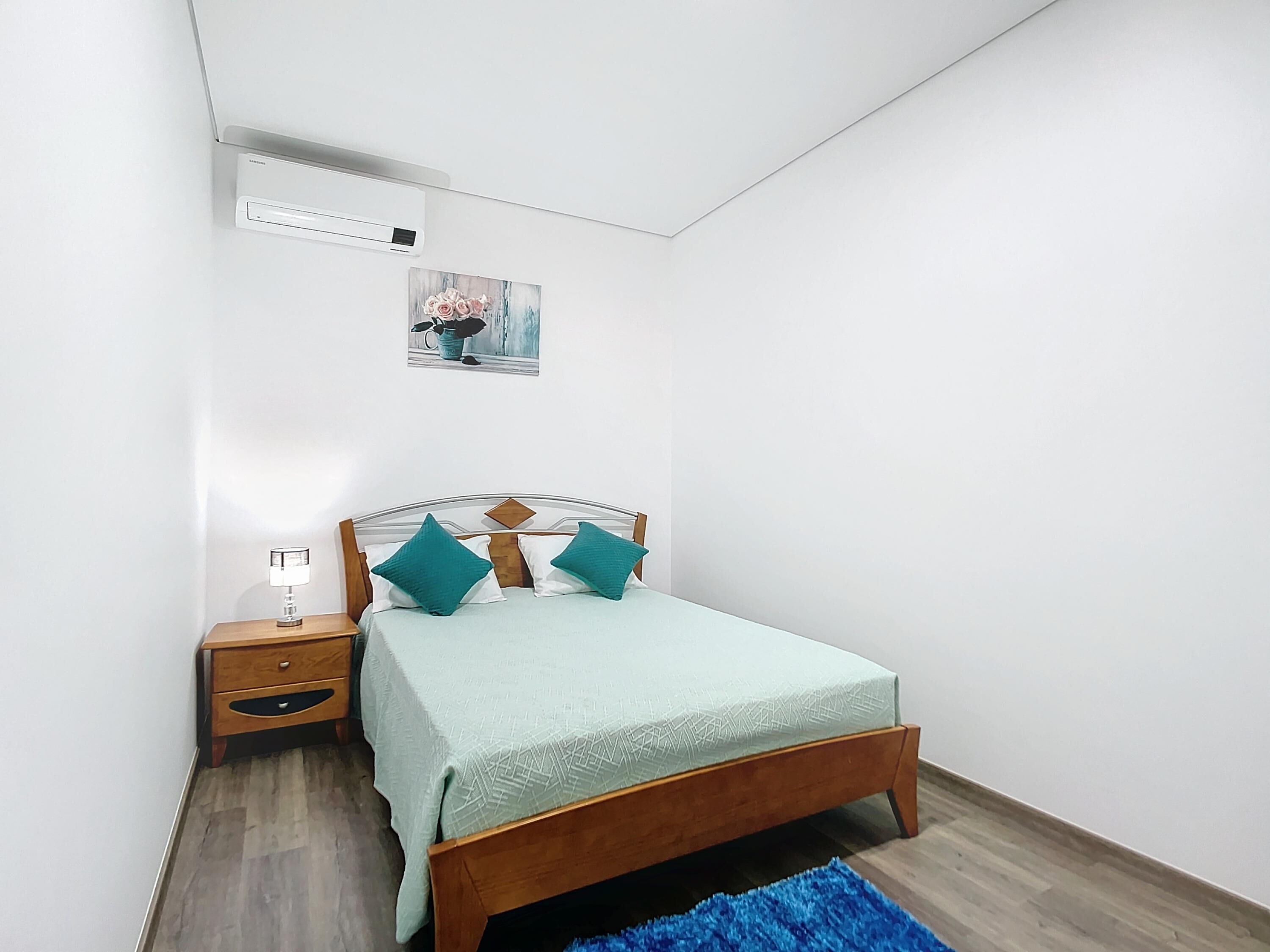 A bedroom with a bed, a nightstand, a lamp, a wall-mounted air conditioner, and a framed picture.