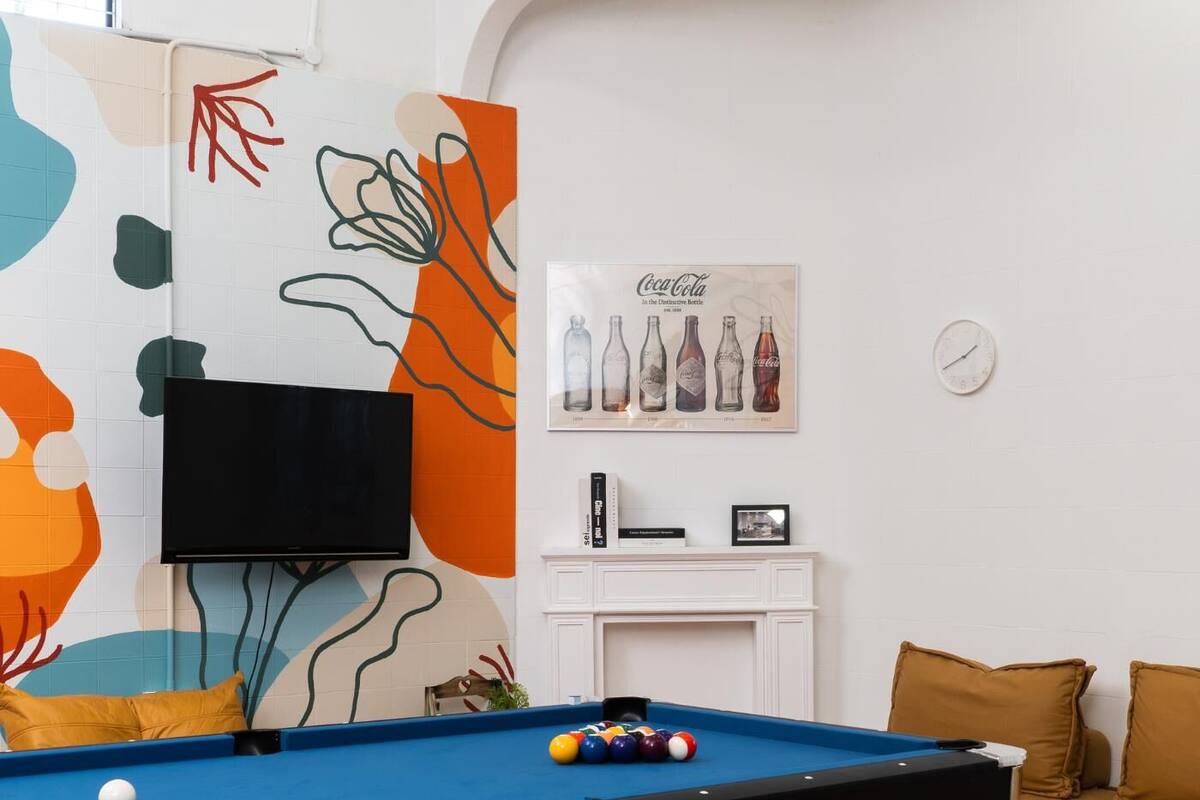 A room with a pool table, a television mounted on the wall, a Coca-Cola vintage bottle poster, and a clock on the wall.
