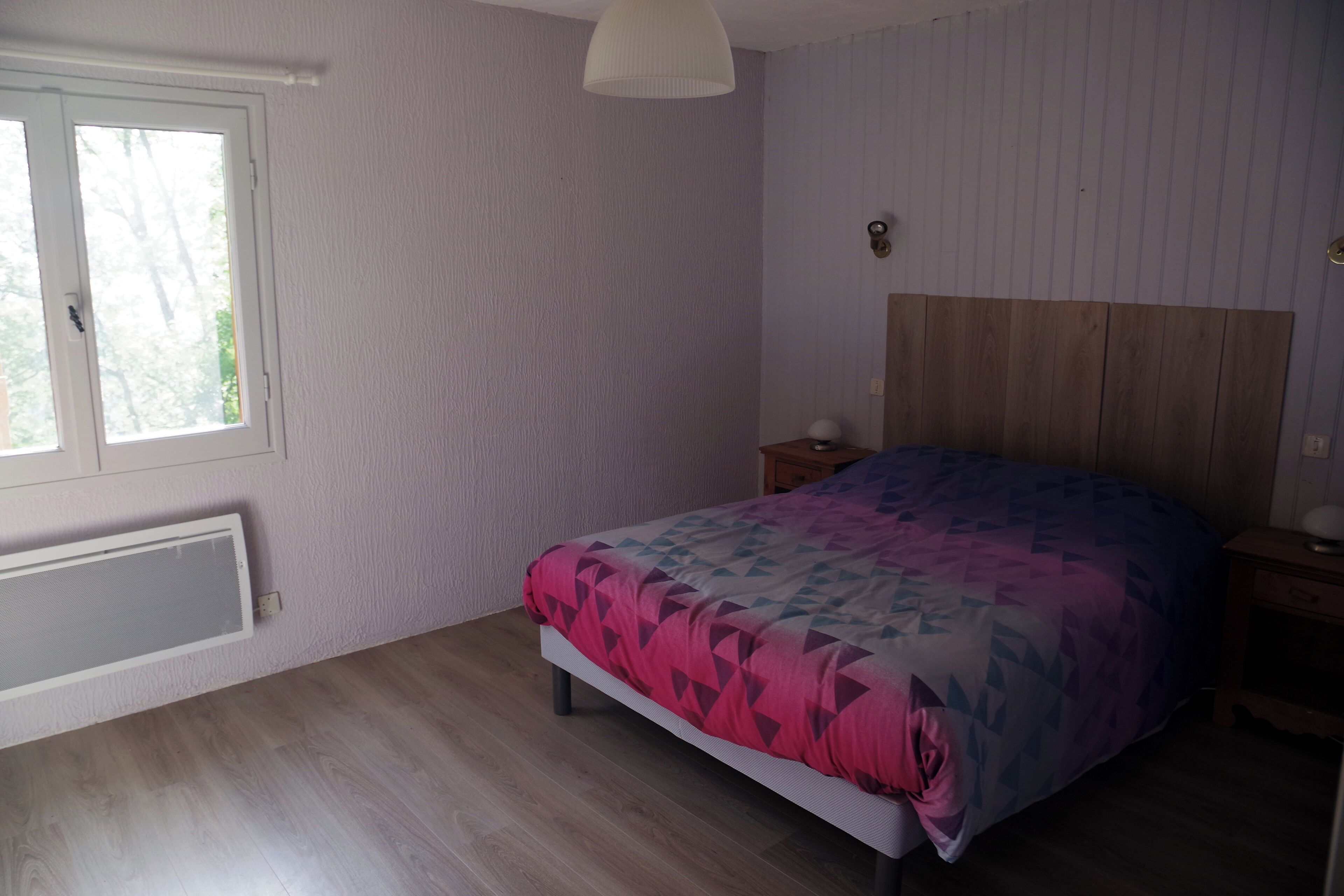 A bedroom with a bed, a nightstand, a wall-mounted lamp, and a window with blinds.
