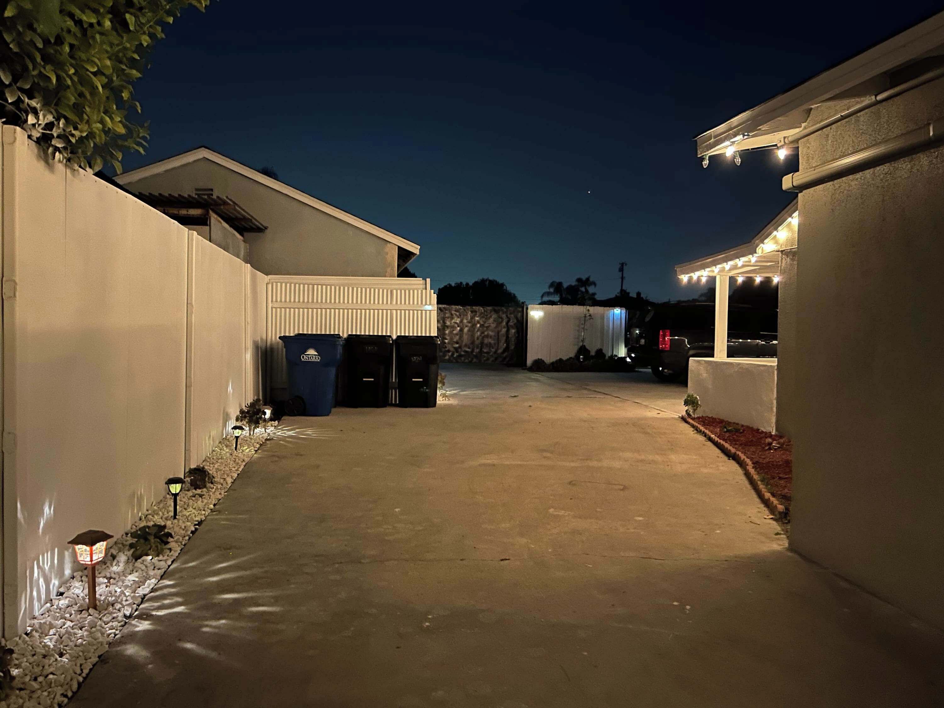 A paved driveway leading to a building with trash bins and a covered area with string lights.
