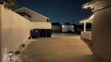 A paved driveway leading to a building with trash bins and a covered area with string lights.