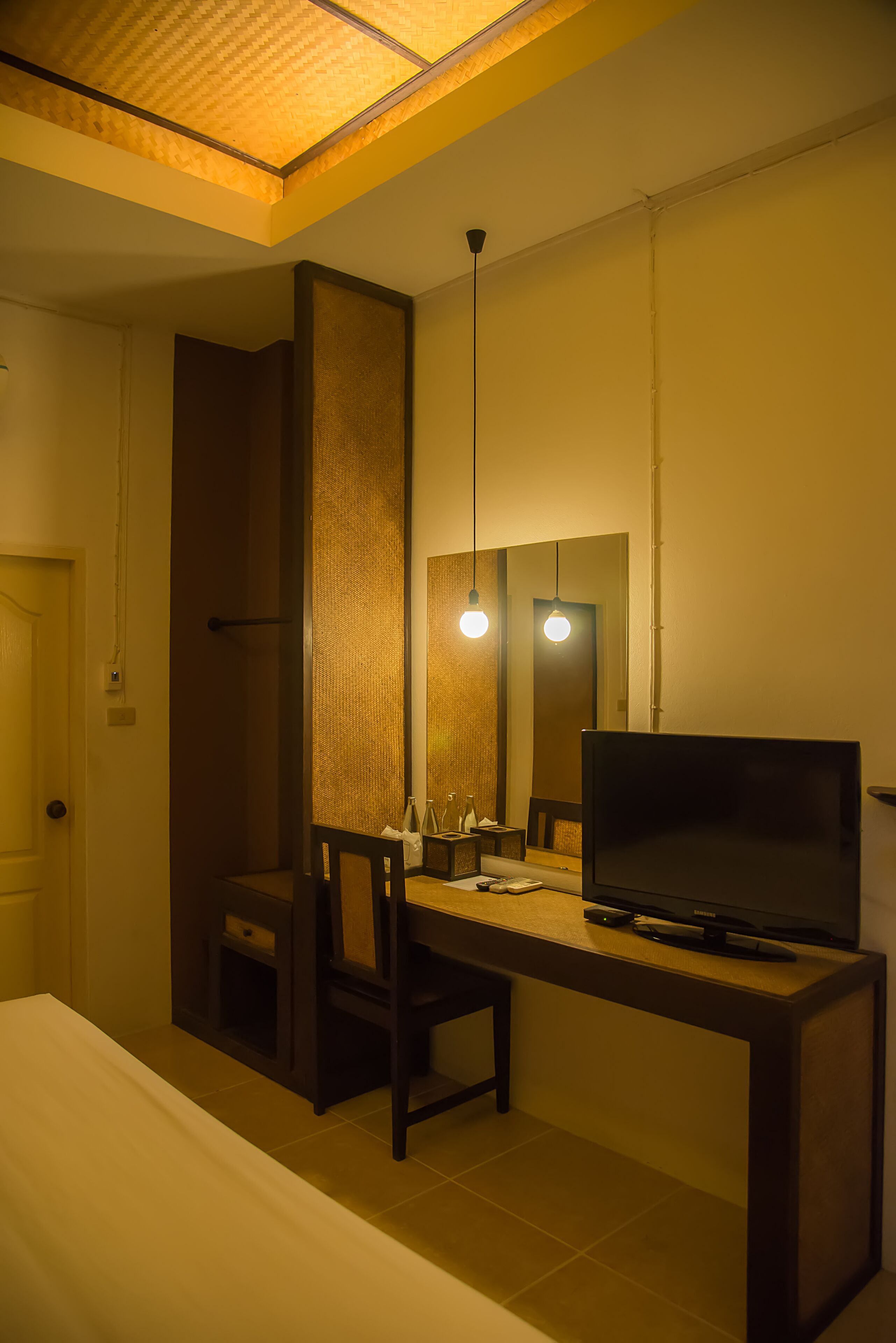 A hotel room with a bed, a desk with a TV, a chair, a mirror, and a hanging light fixture.