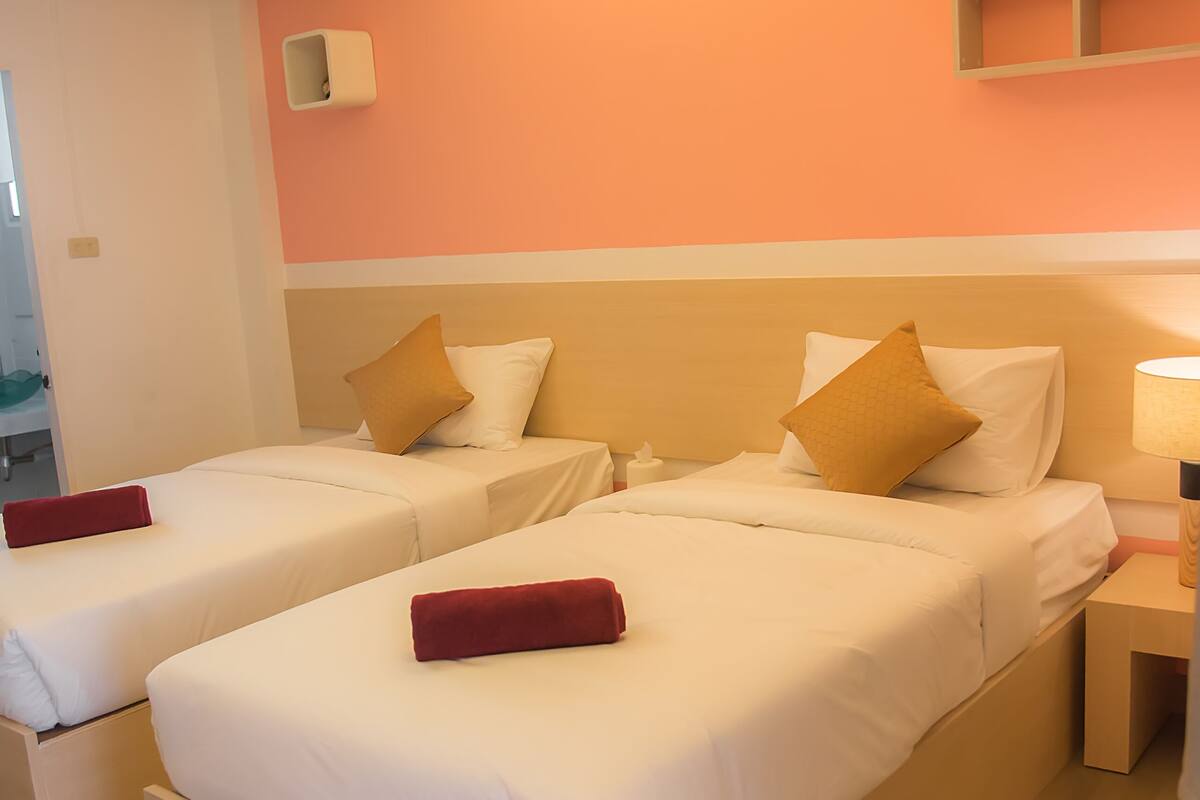 A hotel room with two beds, each with white linens and red pillows. There is a nightstand with a lamp and a small shelf above the beds.