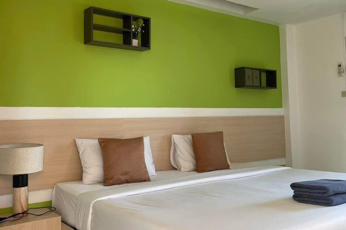 A hotel room with a bed, bedside table, lamp, green wall, and a small shelf with decorative items.