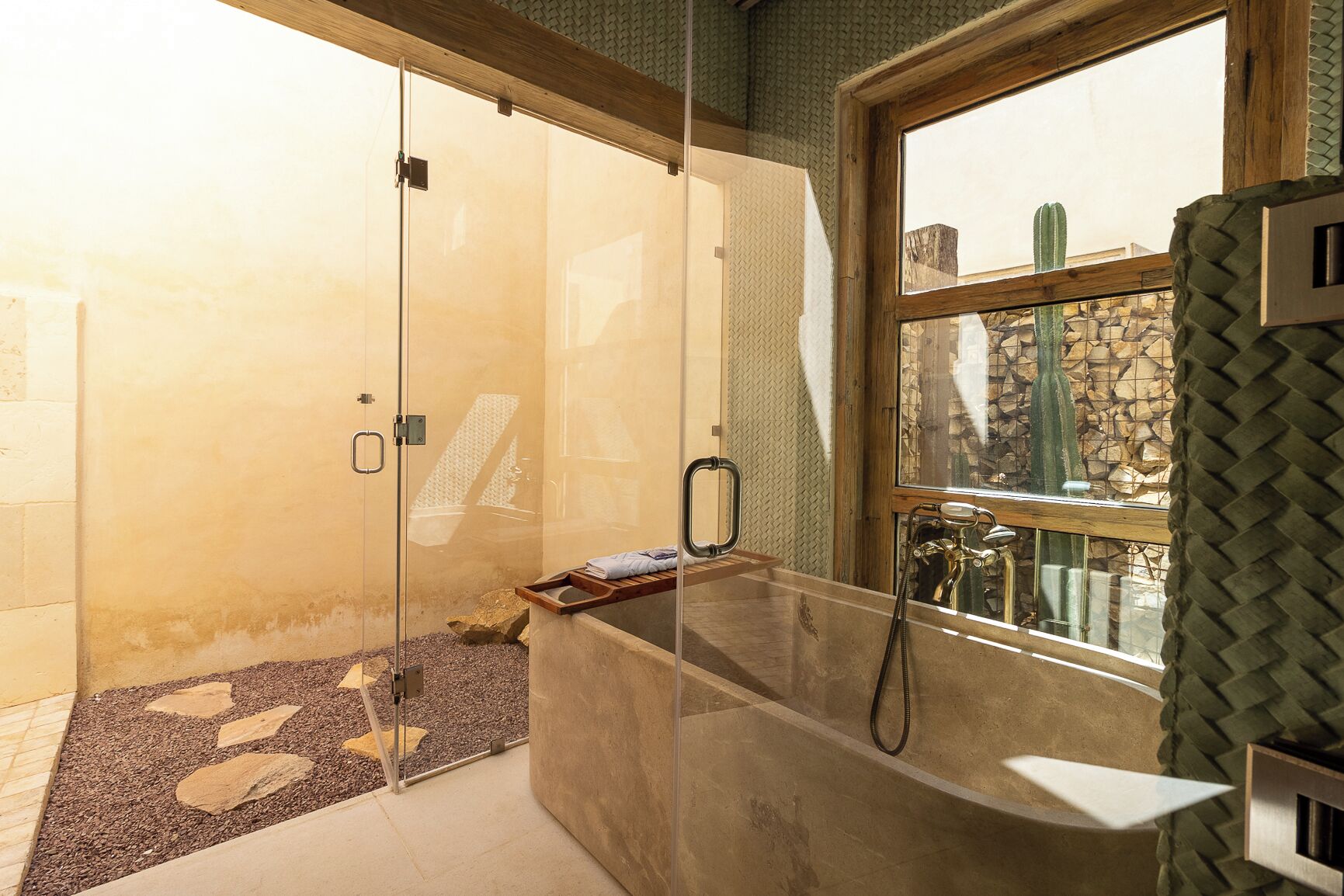 A glass-enclosed shower area with a wooden bench, a stone wall, and a window with a cactus view.