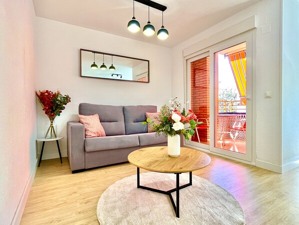 A modern living room with a grey sofa, a round wooden coffee table, a white vase with flowers, and a large mirror on the wall.