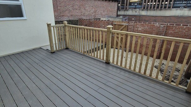 Een houten terras met een metalen hekwerk, een bakstenen gebouw en een heldere lucht.