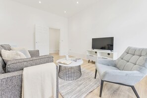 Een moderne woonkamer met een bank, fauteuil, salontafel en een flatscreen televisie op een witte zuil.