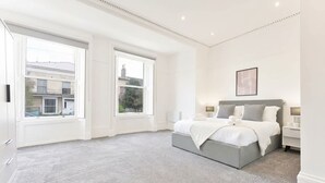Een slaapkamer met een groot raam, een bed met een grijze hoofdbord en een nachtkastje met een lamp.