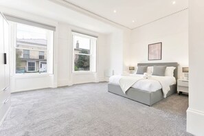 A bedroom with a large window, a bed with a grey headboard, and a bedside table with a lamp. - Spacious 1 Bed Apartment in Cheltenham w Terrace (Cheltenham)