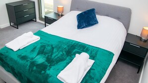 Un lit bien fait, recouvert d’une couverture blanche et turquoise, avec un oreiller bleu et deux serviettes blanches pliées. On aperçoit une petite table de chevet avec une lampe, une commode sombre et une fenêtre donnant sur l’extérieur.