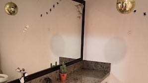 Ein Badezimmer mit einer Granit-Arbeitsplatte, einem weißen Waschbecken und einem Spiegel darüber. Auf der Arbeitsplatte steht ein Blumentopf und über dem Spiegel befindet sich eine Wandleuchte.