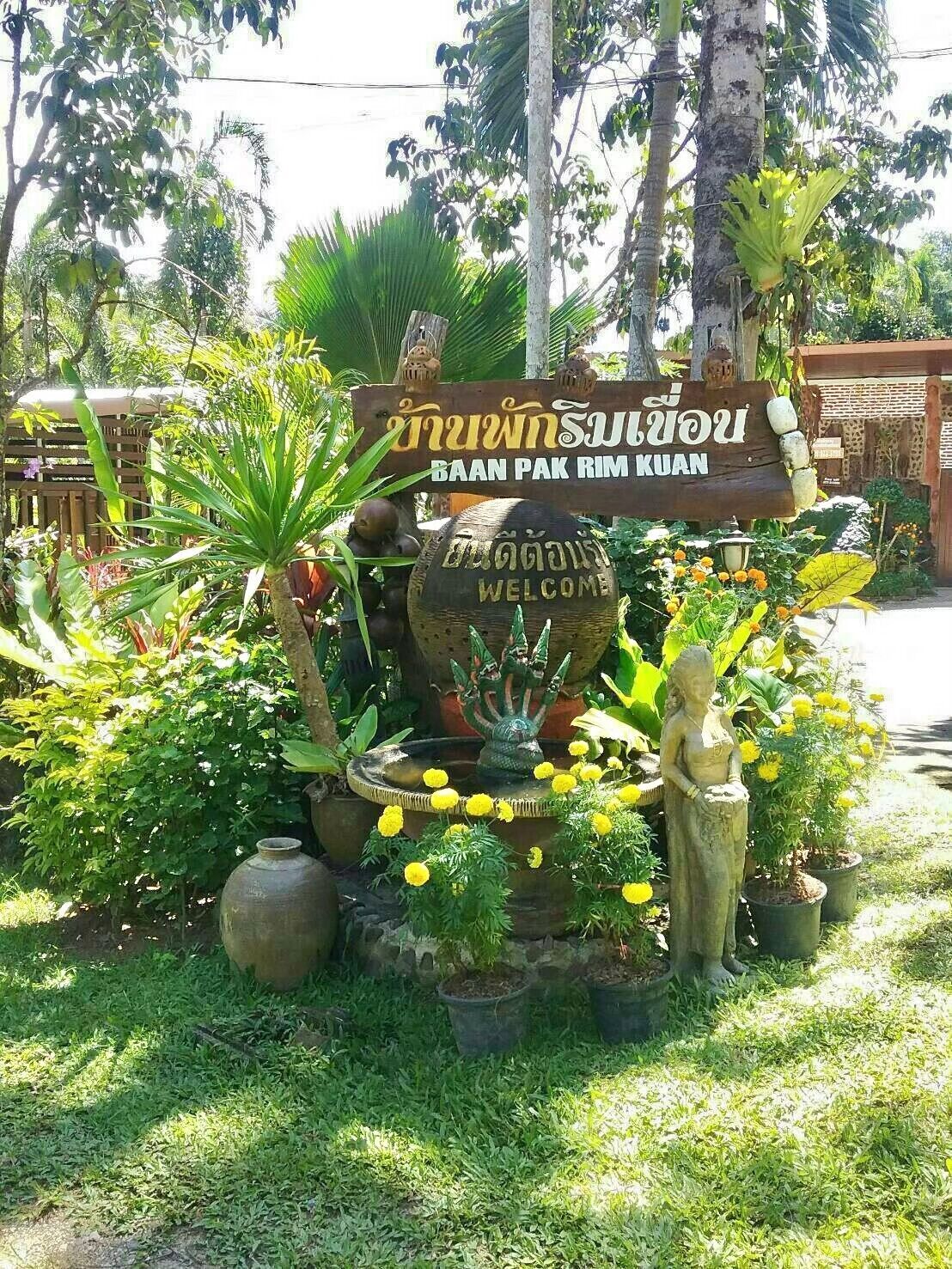 Un letrero de bienvenida con escritura tailandesa, una piedra decorativa con la palabra 'WELCOME' grabada y una estatua de una mujer.