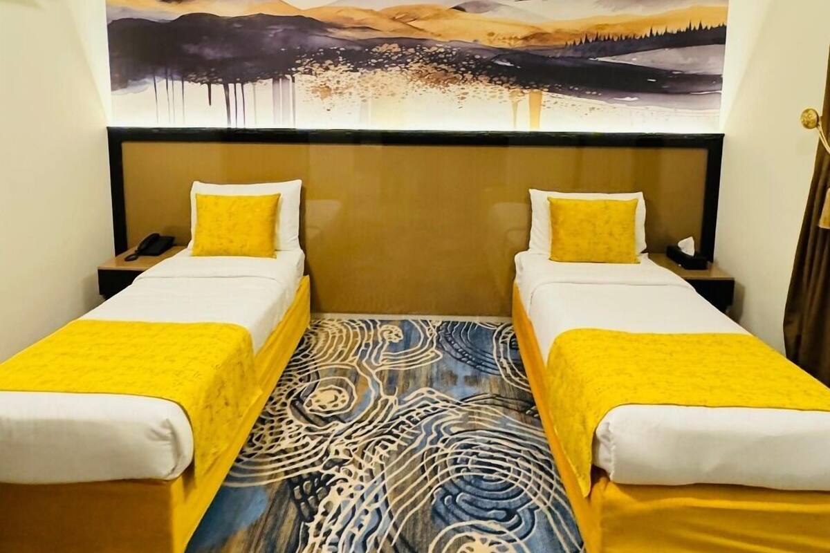 A hotel room with two beds, a large painting of a mountain landscape, and a patterned carpet.