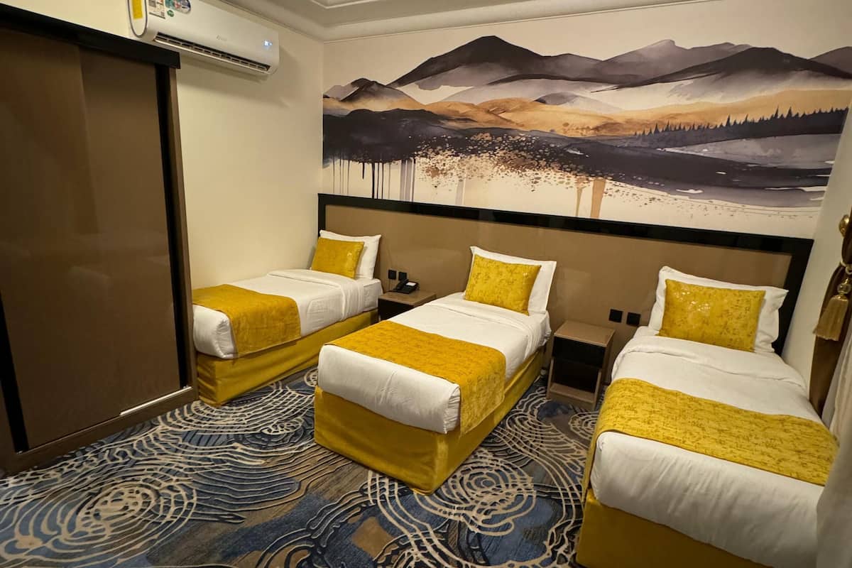 A hotel room with three beds, each with yellow bedding, a large painting of a mountainous landscape on the wall, and a patterned carpet.