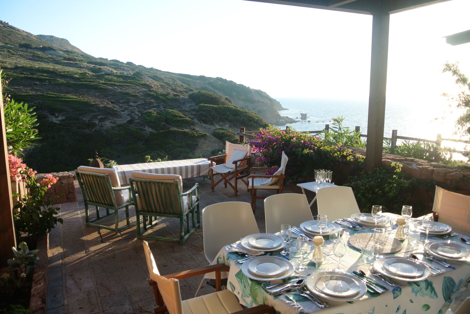 An outdoor dining area with tables set for a meal, overlooking a coastal landscape with hills and the sea.