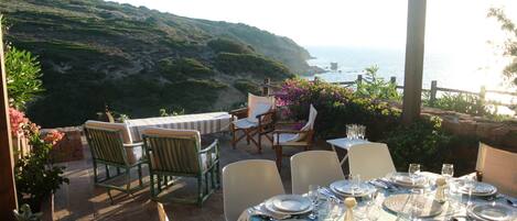An outdoor dining area with tables set for a meal, overlooking a coastal landscape with hills and the sea.