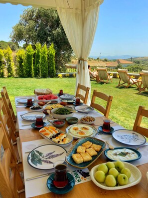 An outdoor dining table set with various dishes and drinks, surrounded by chairs and a scenic view of trees and hills. - Vinea Bozcaada (Bozcaada)