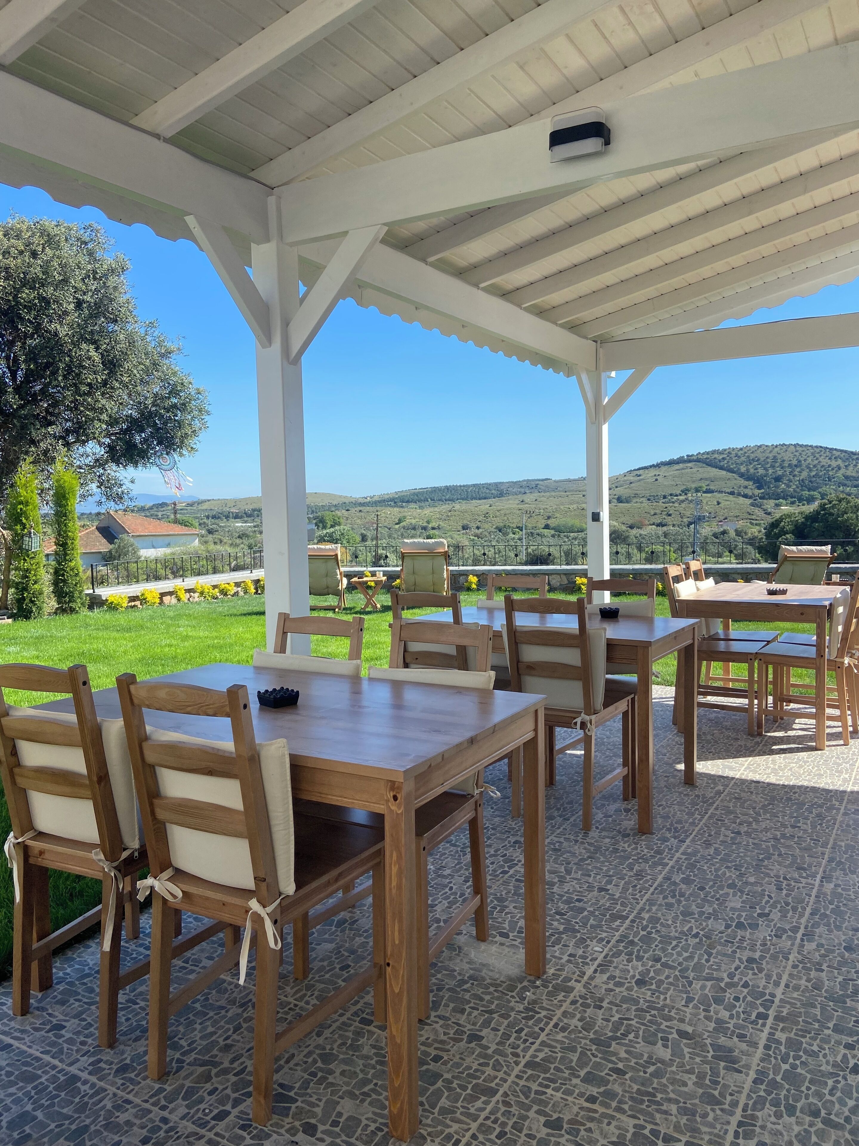 An outdoor patio with wooden tables and chairs, a white ceiling, and a view of green hills and a tree.