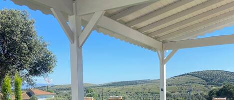 An outdoor patio with wooden tables and chairs, a white ceiling, and a view of green hills and a tree.