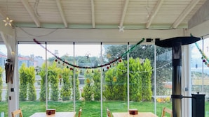 Eine Terrasse mit Holztisch und -stühlen, einem schwarzen Grill und Blick auf einen grünen Garten durch große Fenster.
