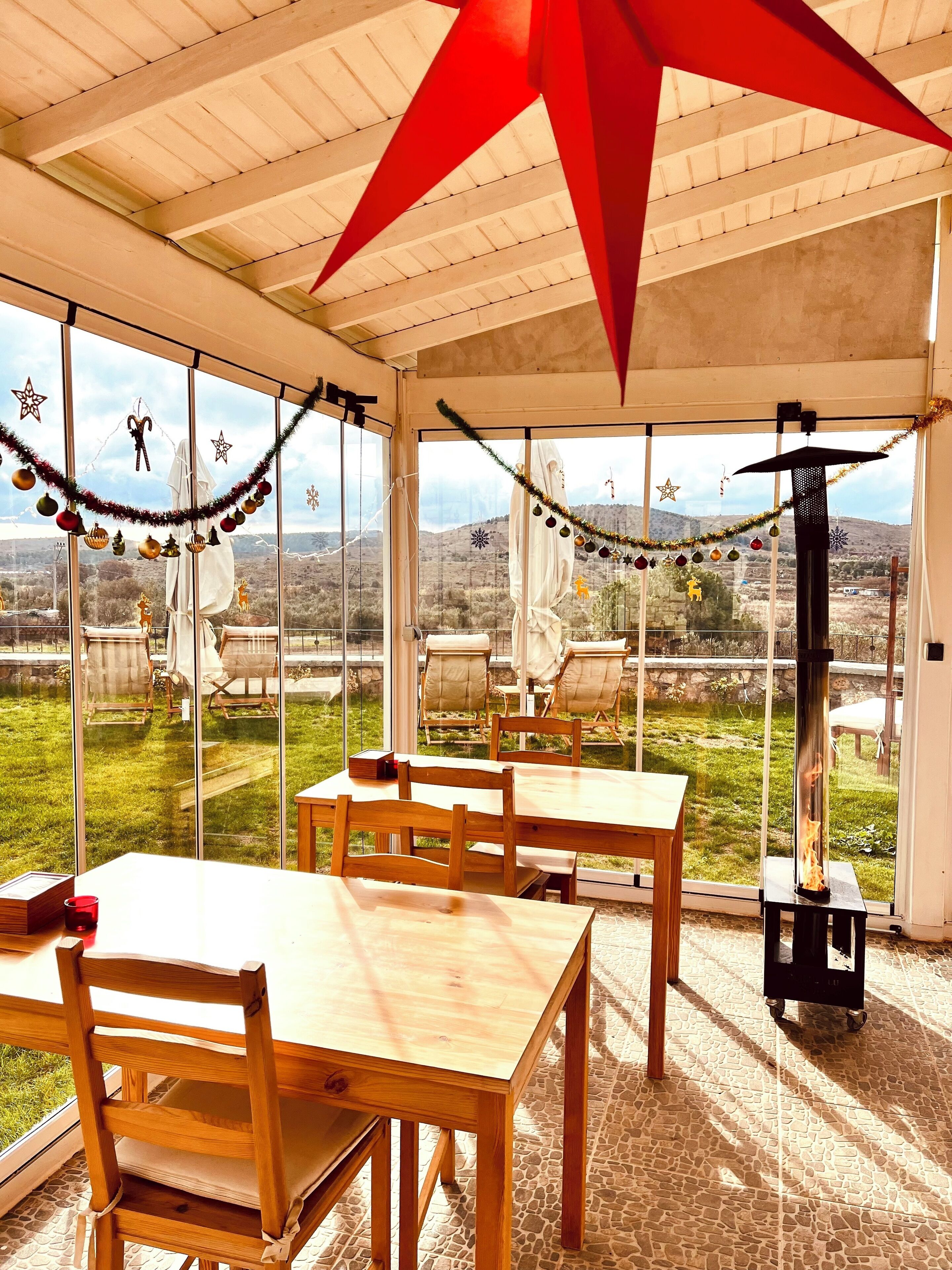 A covered outdoor seating area with wooden tables and chairs, decorated with festive ornaments, overlooking a grassy landscape with distant hills.