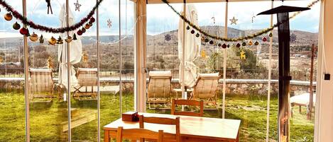 A covered outdoor seating area with wooden tables and chairs, decorated with festive ornaments, overlooking a grassy landscape with distant hills.