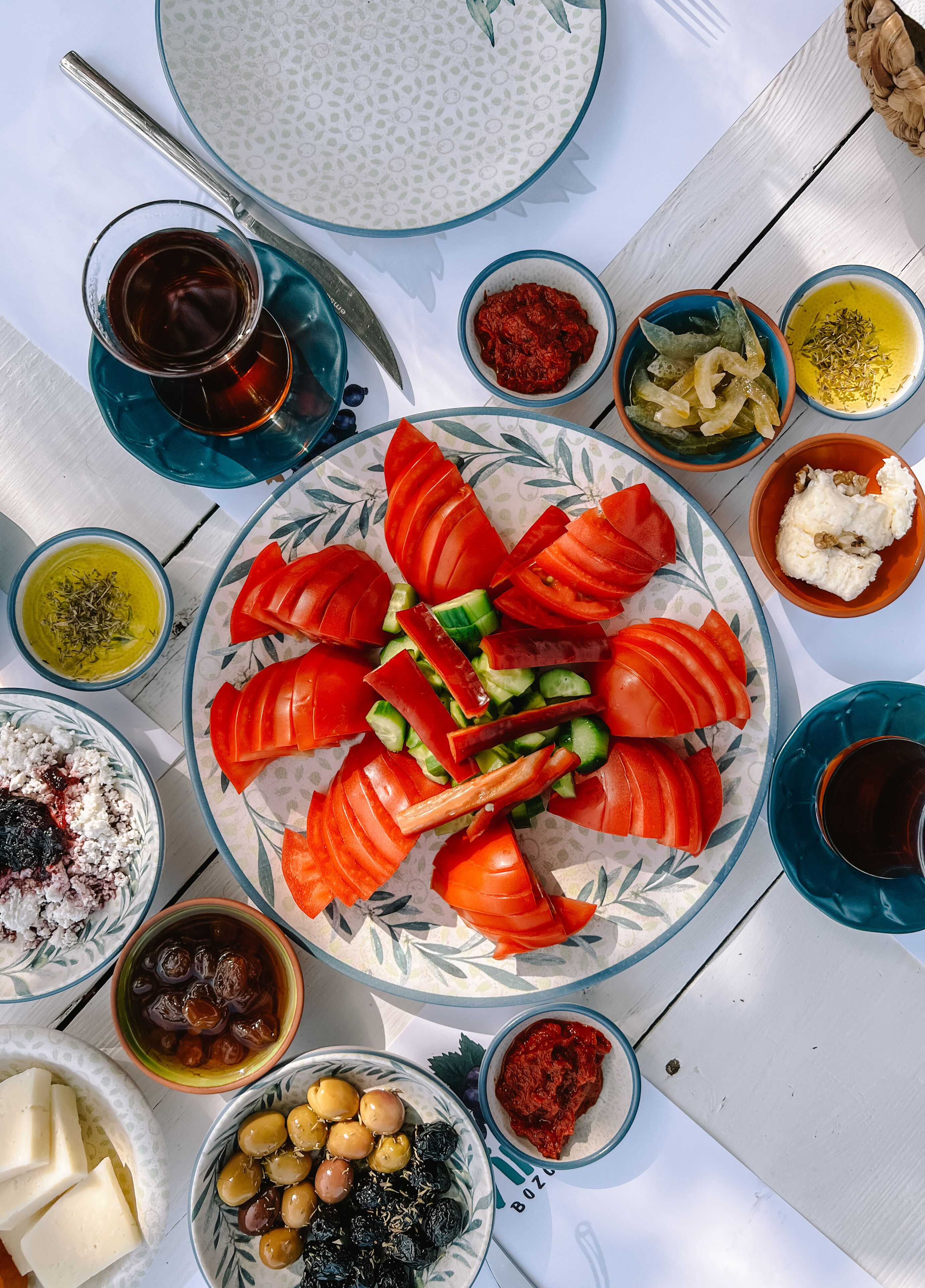 A table with a variety of dishes including sliced tomatoes, cucumbers, olives, and a bowl of yogurt.