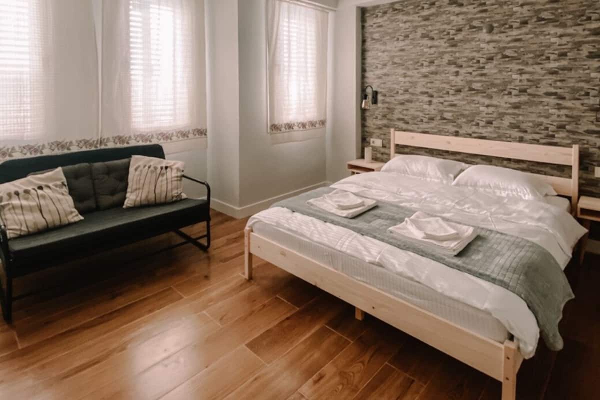 A bedroom with a wooden floor, a bed with white bedding, a dark sofa with patterned cushions, and a stone-textured wall.