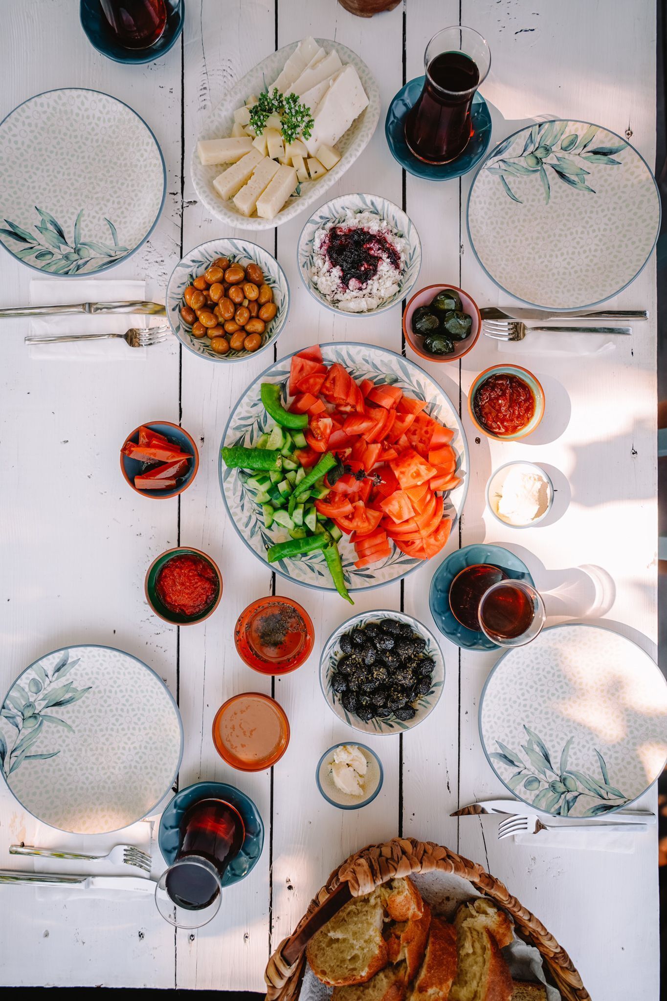 Una mesa con diversos platos, entre ellos una ensalada, pan y recipientes con aceitunas y tomates, sobre una mesa de madera blanca.