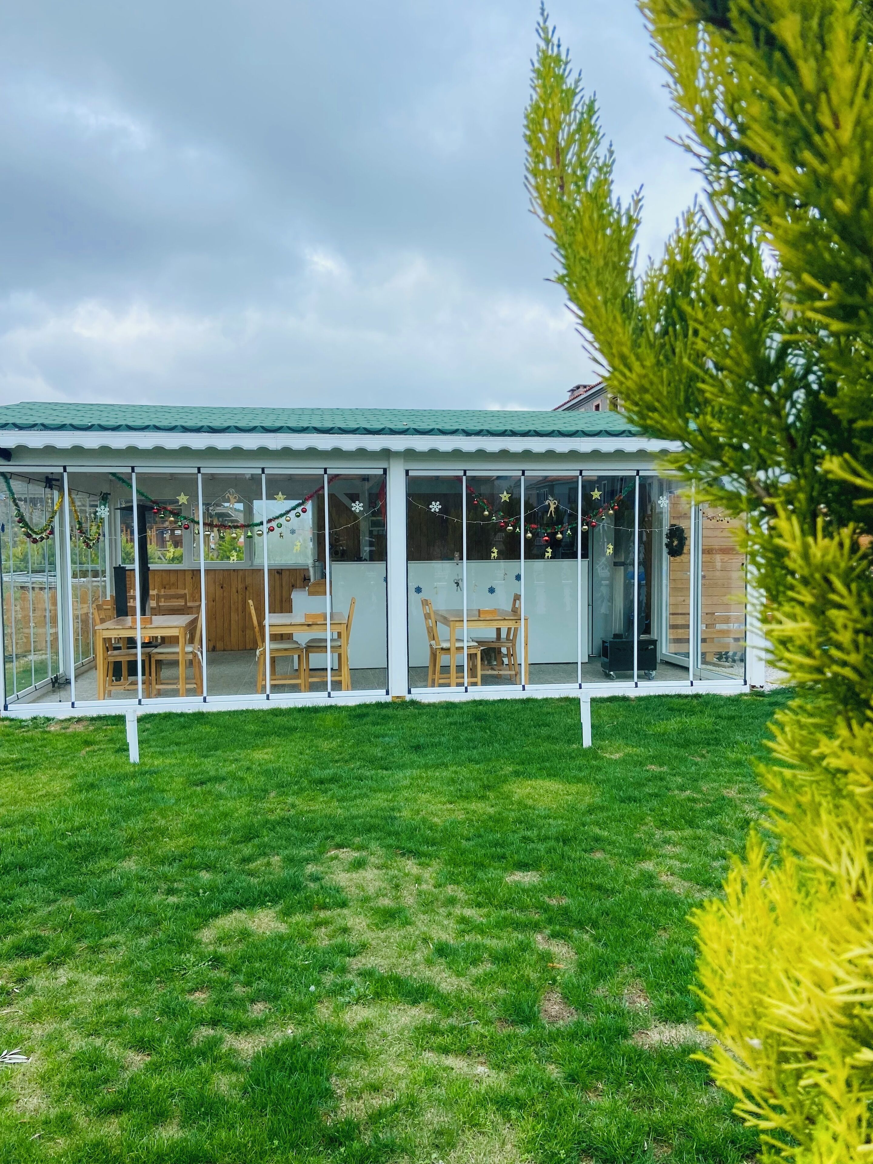 A covered patio area with wooden furniture and glass walls, surrounded by a grassy lawn.