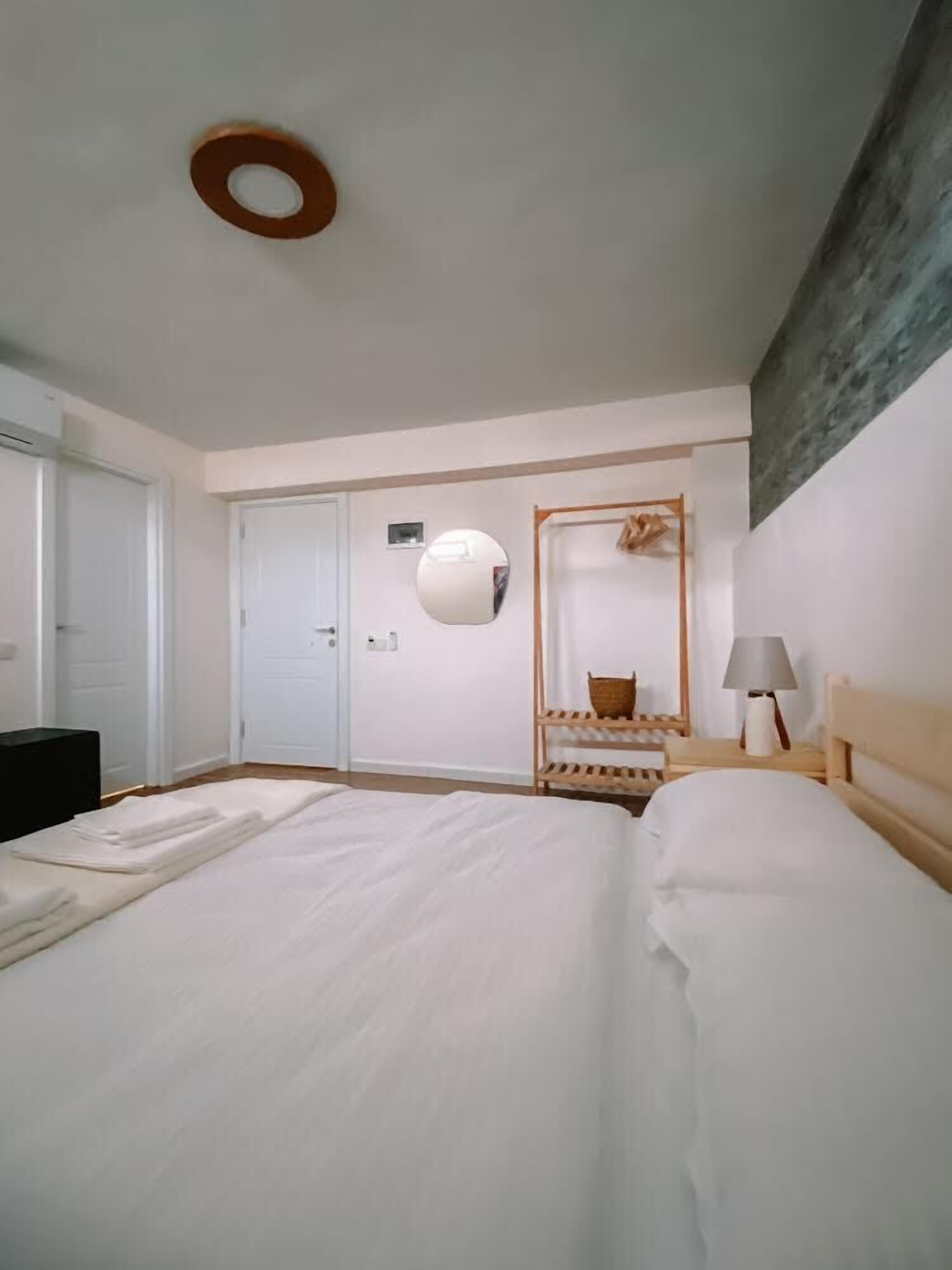 Habitación de hotel con una cama grande, una mesita de noche de madera con una lámpara, un espejo y un pequeño estante de madera con una cesta.