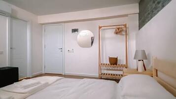 Habitación de hotel con una cama grande, una mesita de noche de madera con una lámpara, un espejo y un pequeño estante de madera con una cesta.