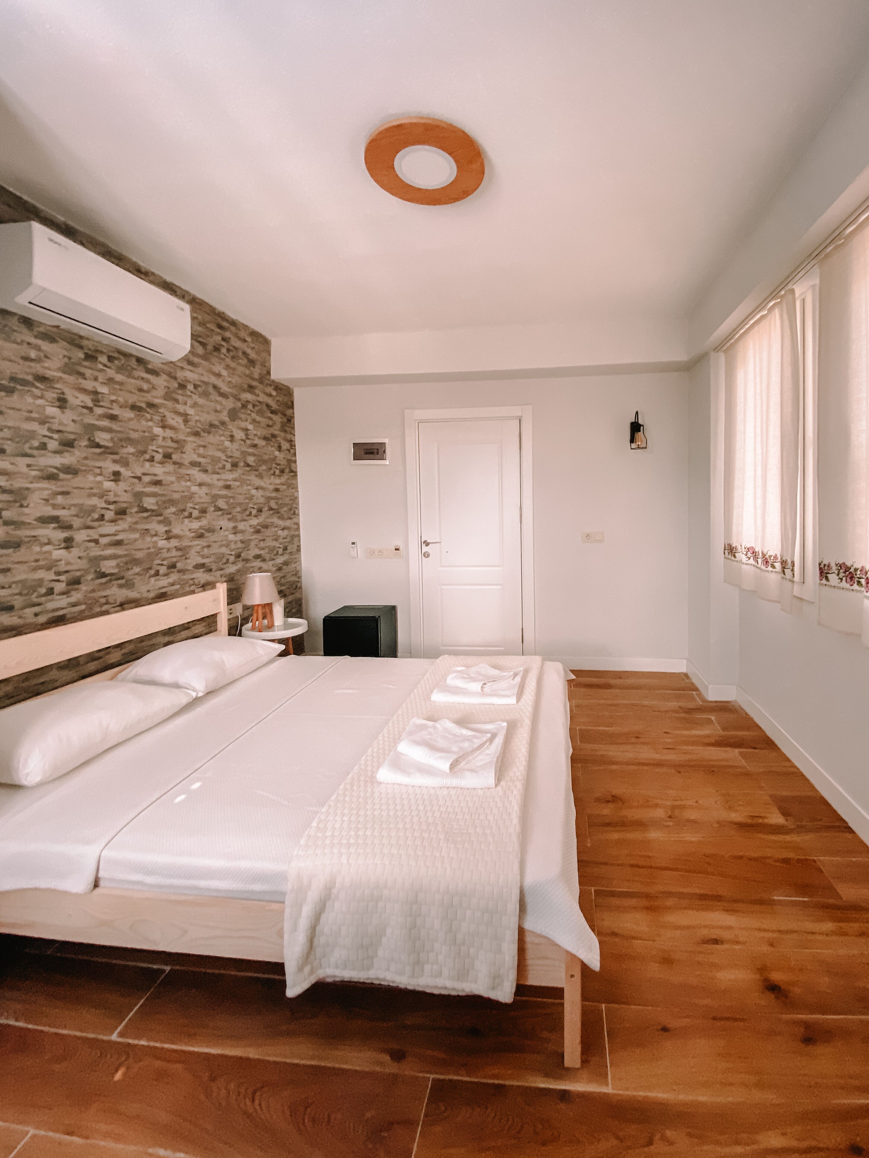 A bedroom with a bed, a wall-mounted air conditioner, a bedside table with a lamp, a door, and a window with curtains.