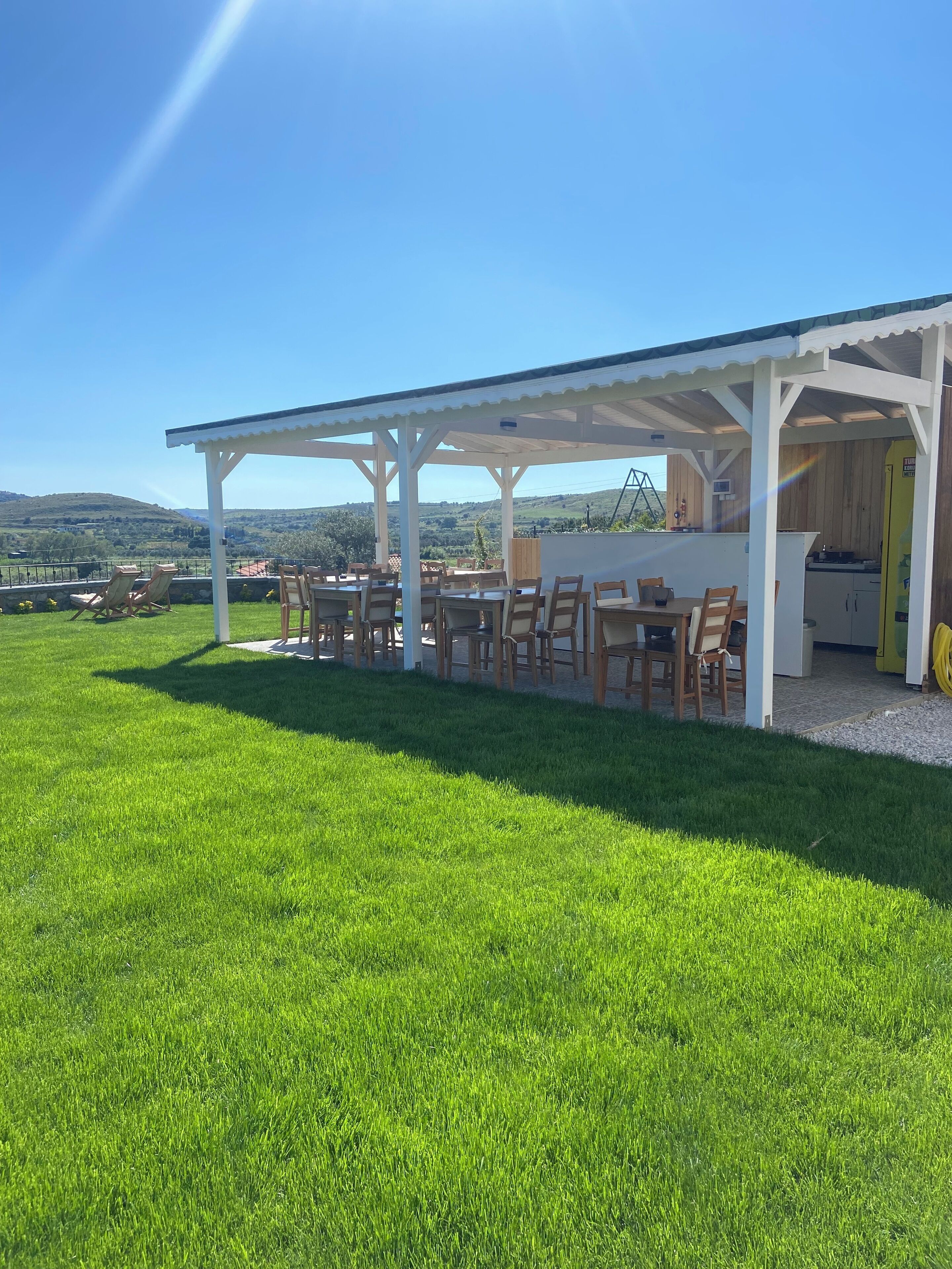 A covered outdoor seating area with tables and chairs, a kitchen area, and a view of the surrounding landscape.