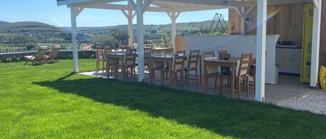 A covered outdoor seating area with tables and chairs, a kitchen area, and a view of the surrounding landscape.